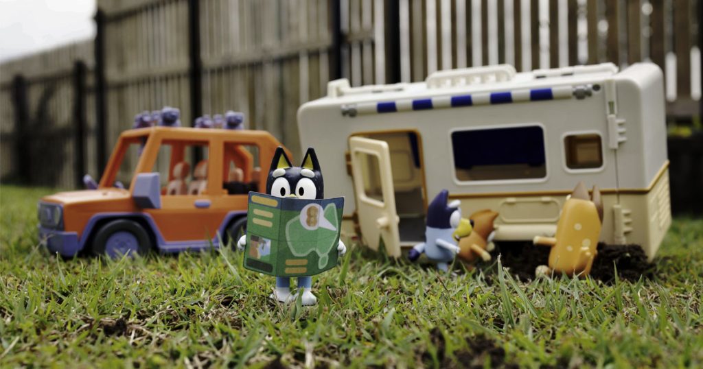 Lifestyle photography of the toy set with Heeler family figurines on some grass with the toy caravan and toy 4-Wheel Drive car in the background.