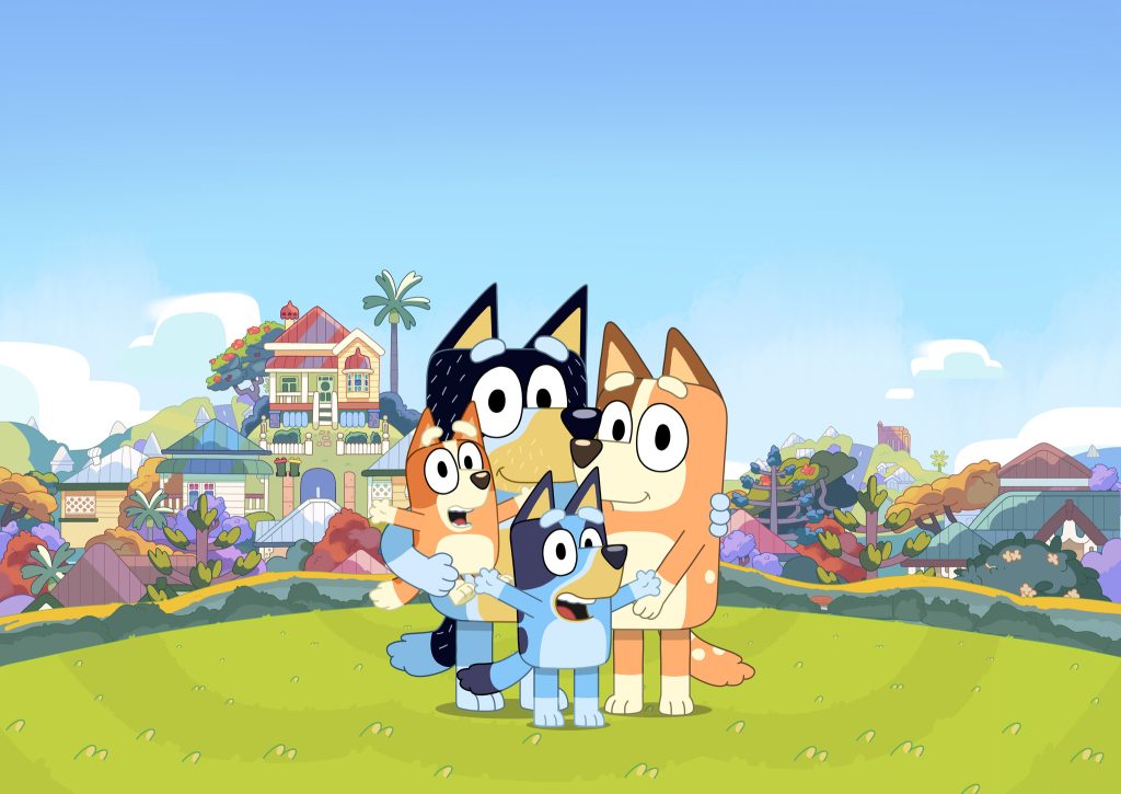 Bluey, Bingo, Mum and Dad stand together smiling in a bright outdoor setting with a clear blue sky.